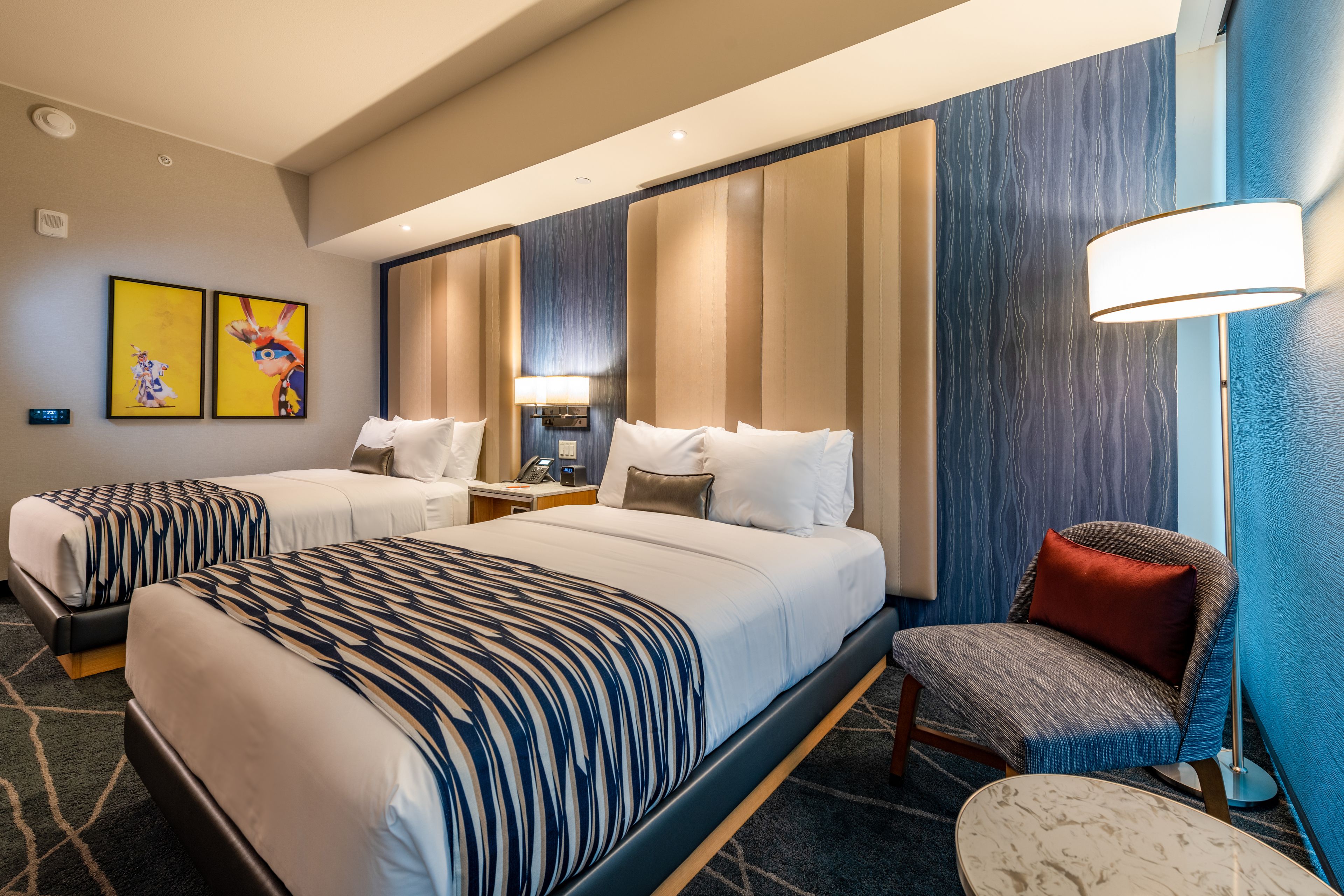 A hotel room with two beds, a chair, a desk, and a lamp.
