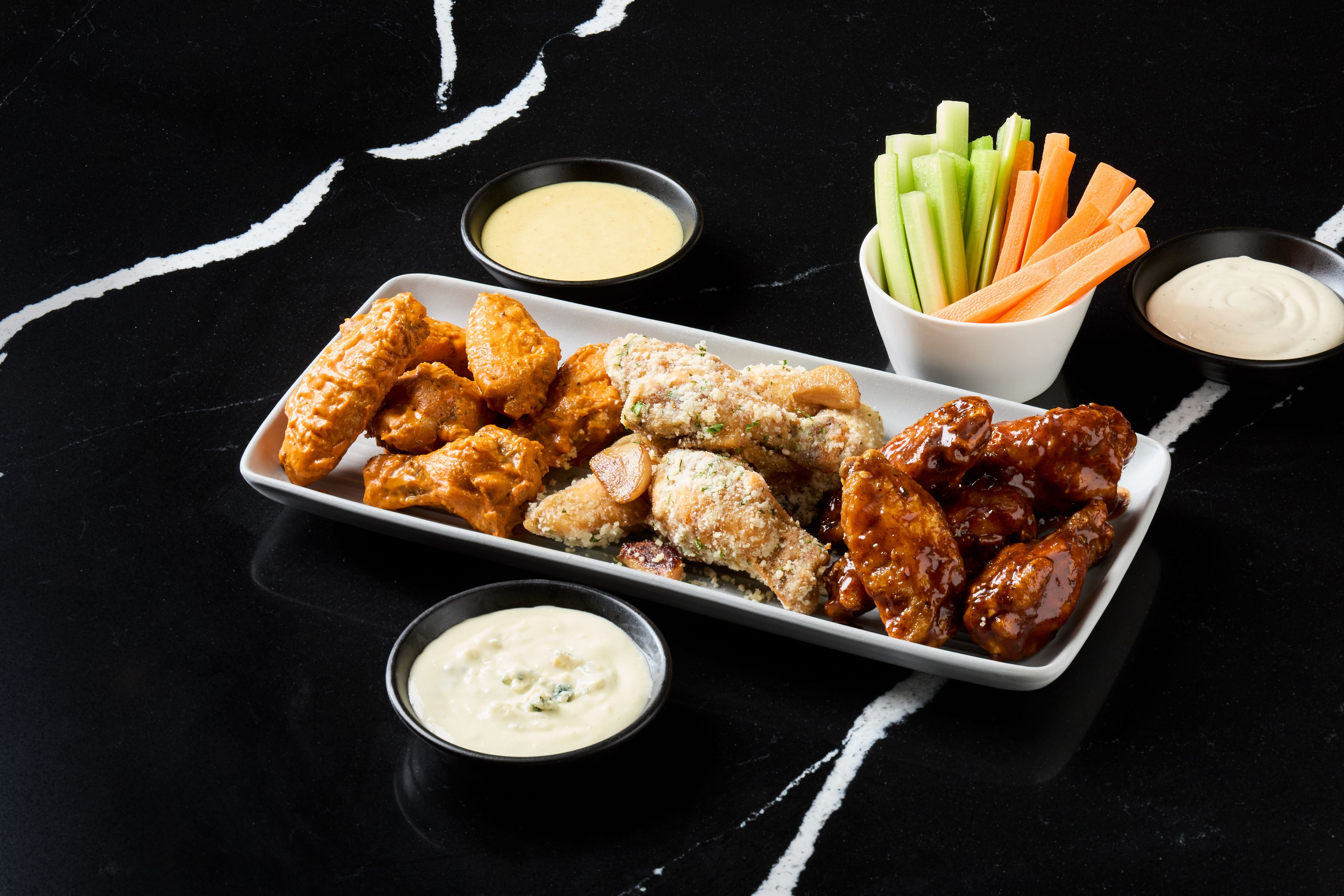 Una bandeja de alitas de pollo con diferentes salsas para mojar, palitos de apio y zanahoria.