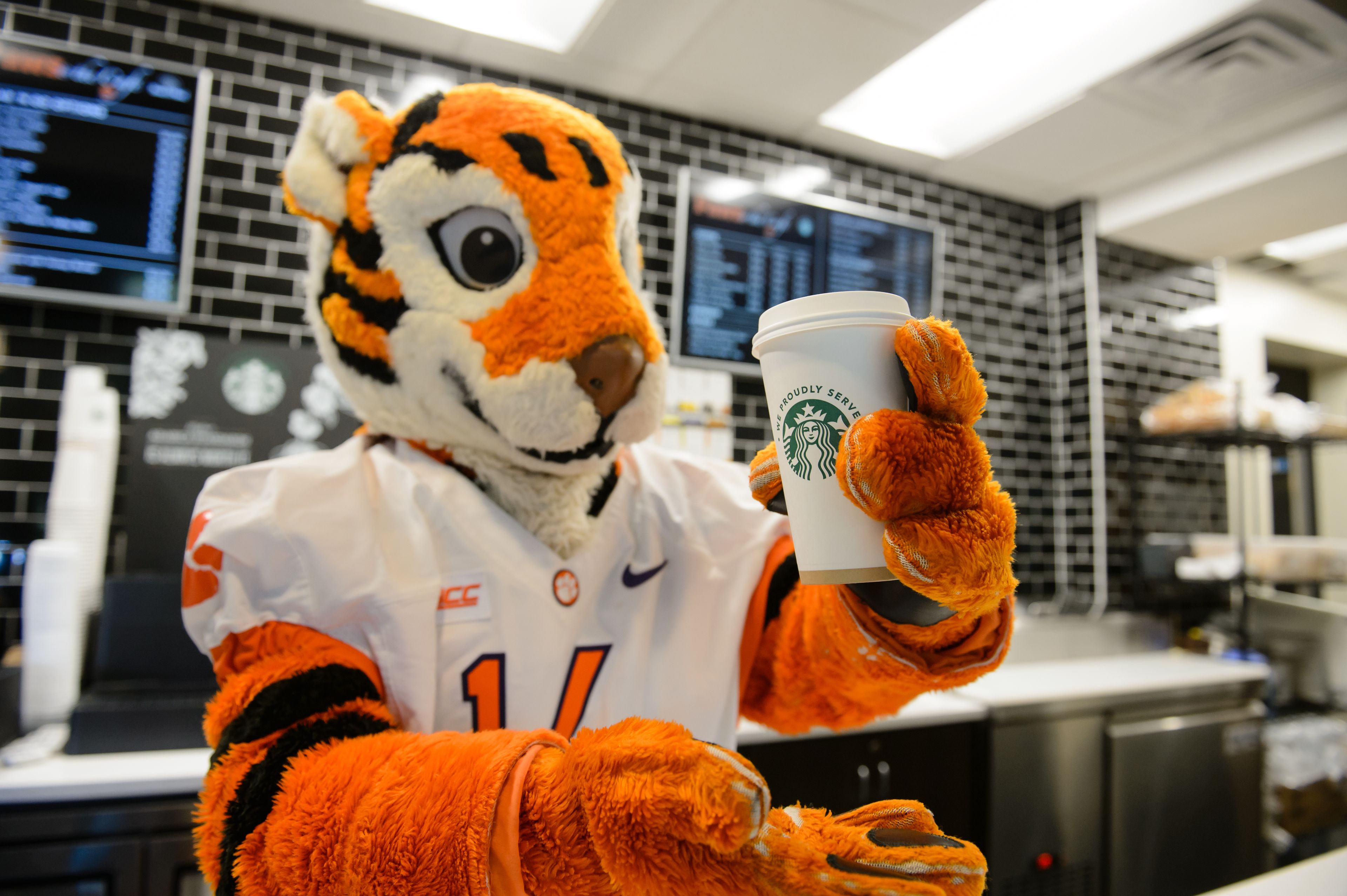 Ein Maskottchen hält eine Starbucks-Tasse in einem modernen, hell erleuchteten Coffeeshop mit digitalen Menütafeln.