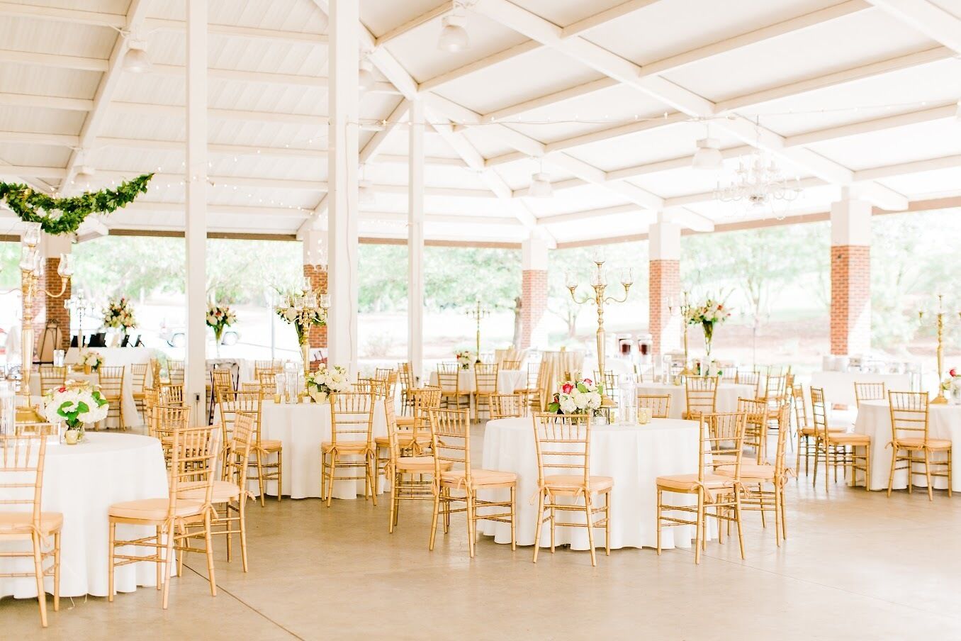 A spacious indoor venue with high ceilings and large windows, featuring multiple round tables with white tablecloths and gold chairs, arranged in a spacious layout.