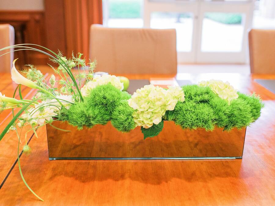 Un jarrón de cristal rectangular con musgo verde y flores blancas sobre una mesa de madera.