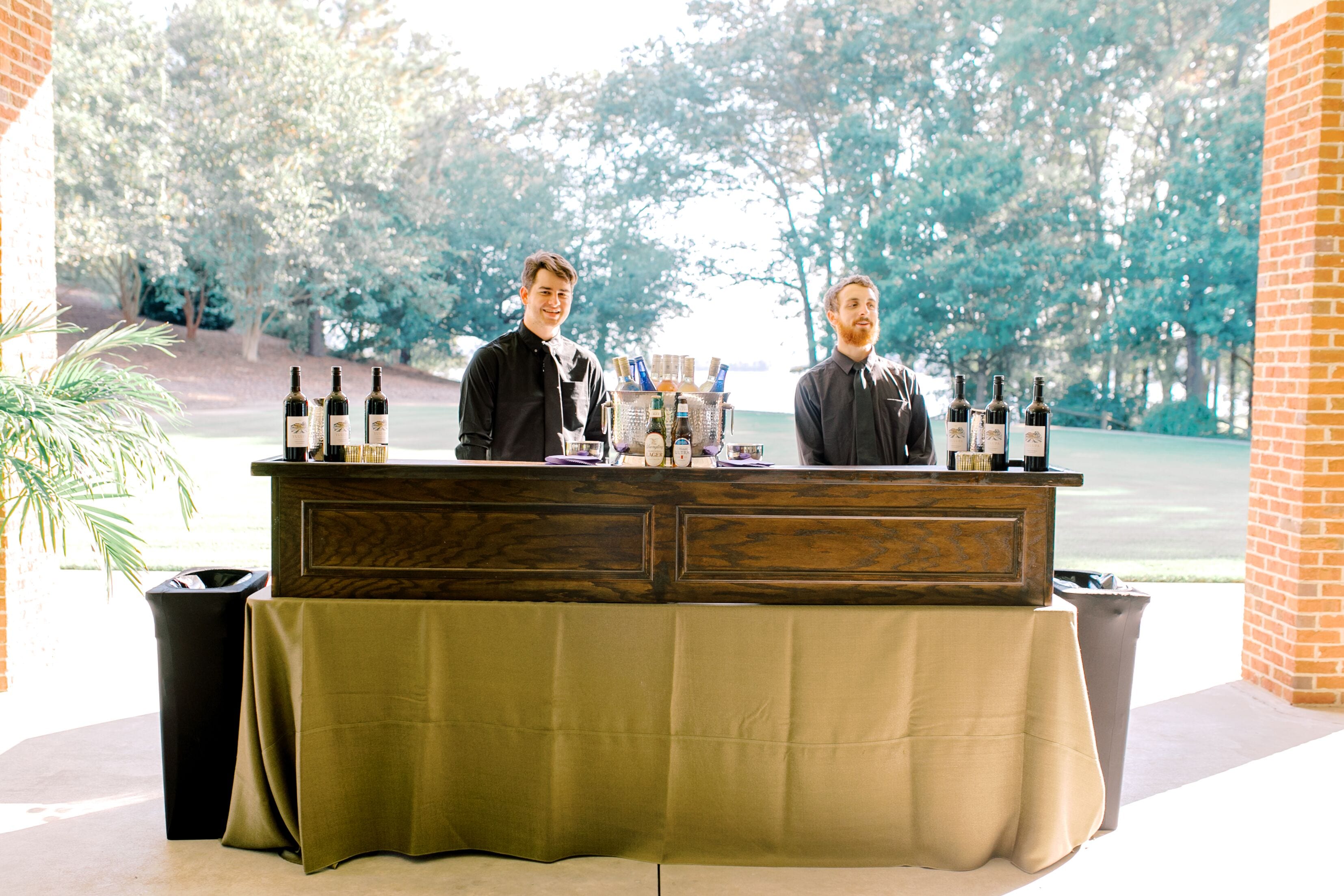 A bar area with two bartenders, a wooden bar counter, and a view of trees and a building in the background.