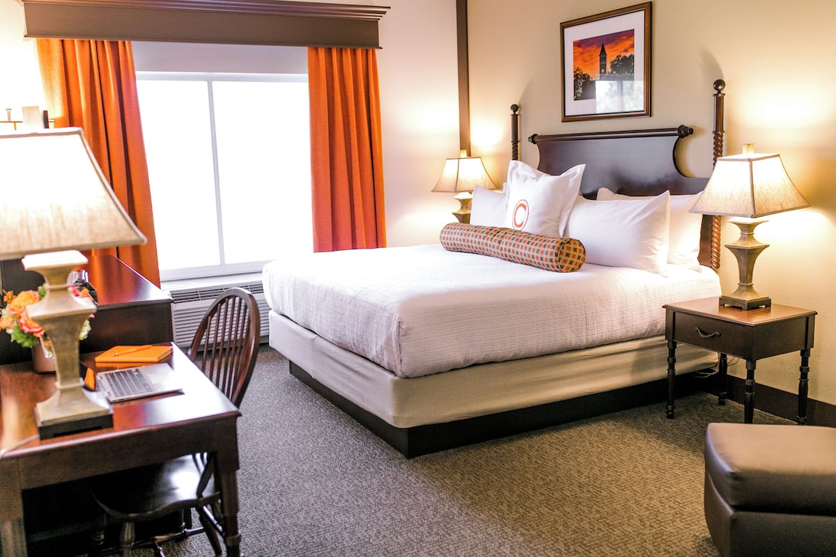 A hotel room with a bed, two bedside tables with lamps, a desk with a chair, and a window with curtains.