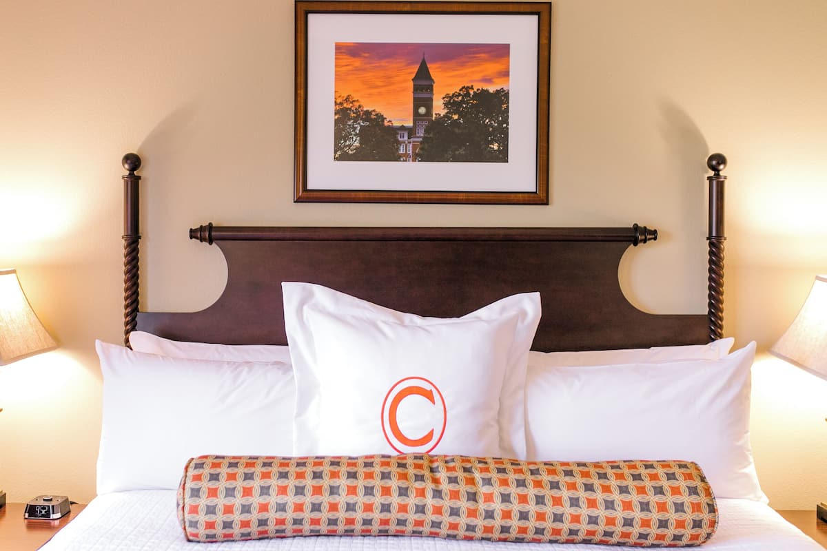 A neatly made bed with a patterned throw, a pillow with a large 'C', and a framed picture of a tower on the wall.