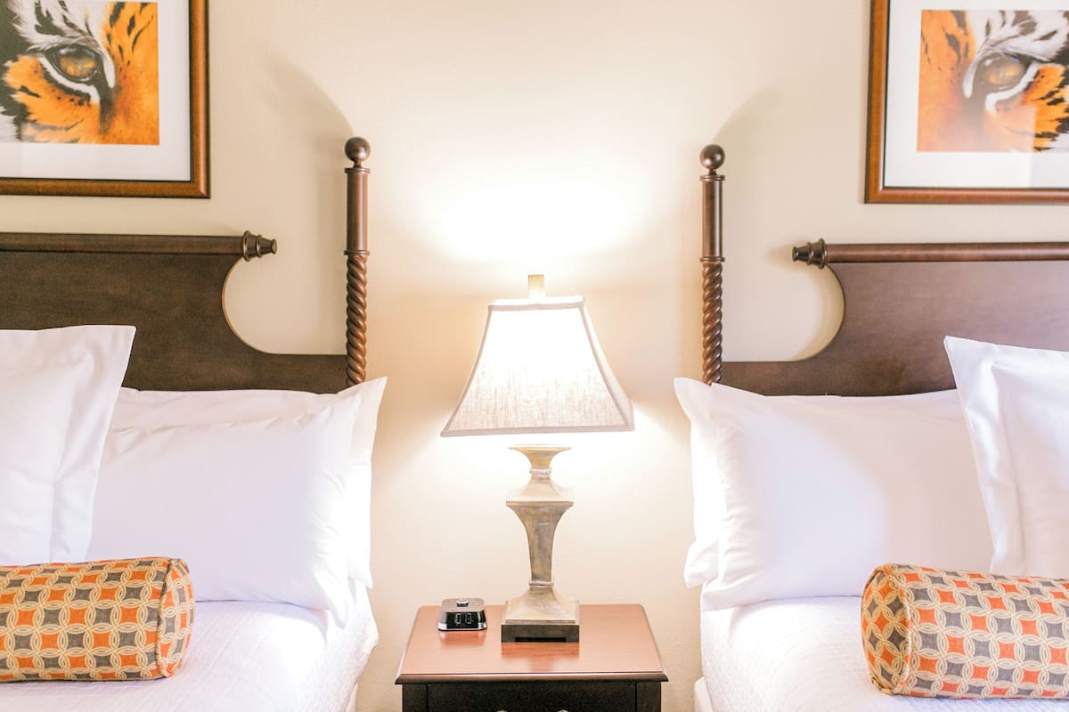 A hotel room with two beds, a nightstand with a lamp, a clock, and framed artwork on the walls.
