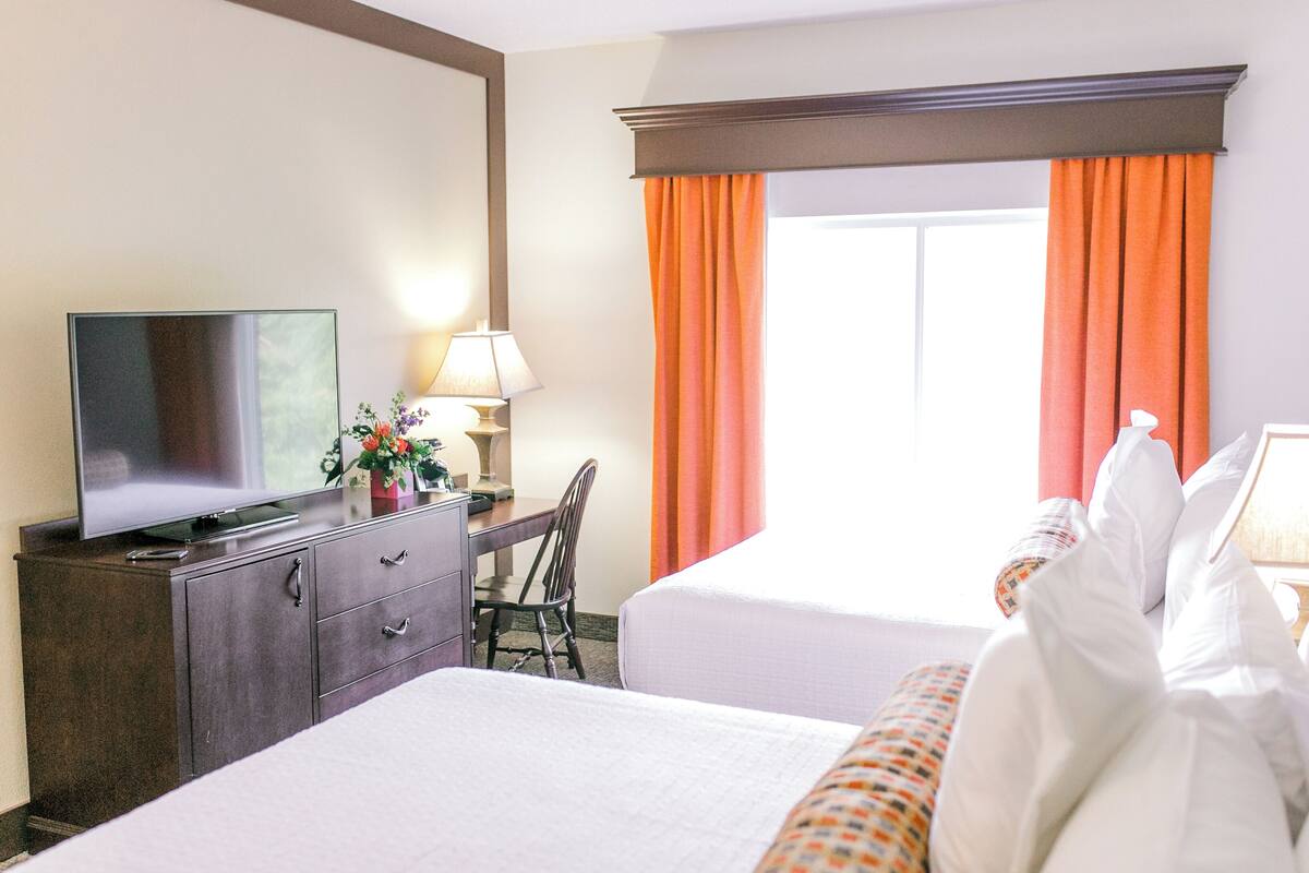 A hotel room with two beds, a TV on a dresser, a desk with a lamp, and a window with orange curtains.