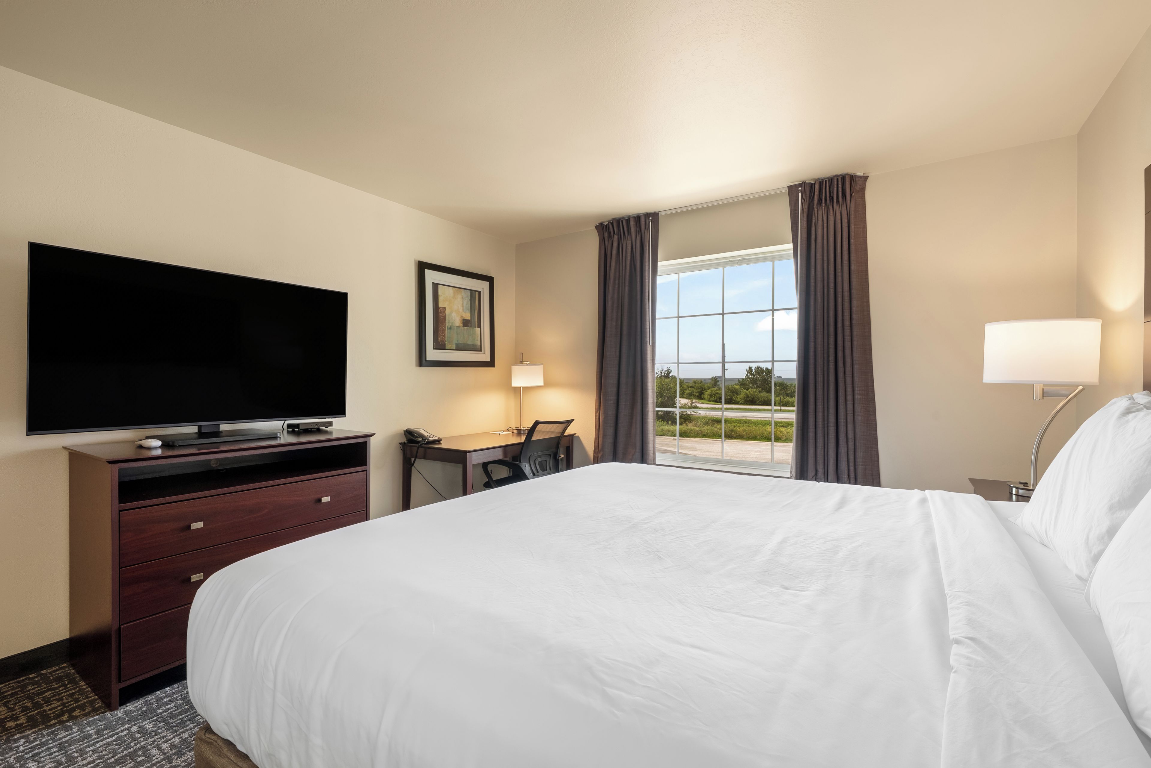 A hotel room with a bed, a desk with a chair, a television, a lamp, and a window with curtains.
