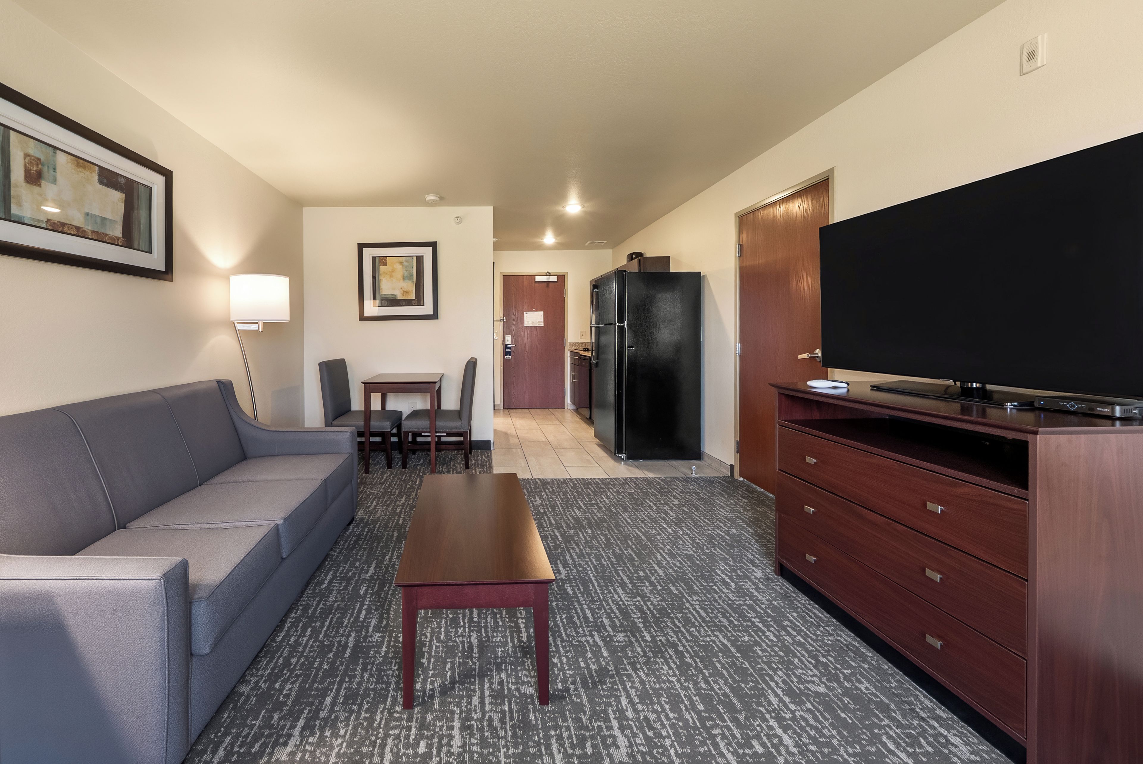 Habitación de hotel con sofá, una mesa, un televisor, un refrigerador y una puerta que da a una zona de cocina.