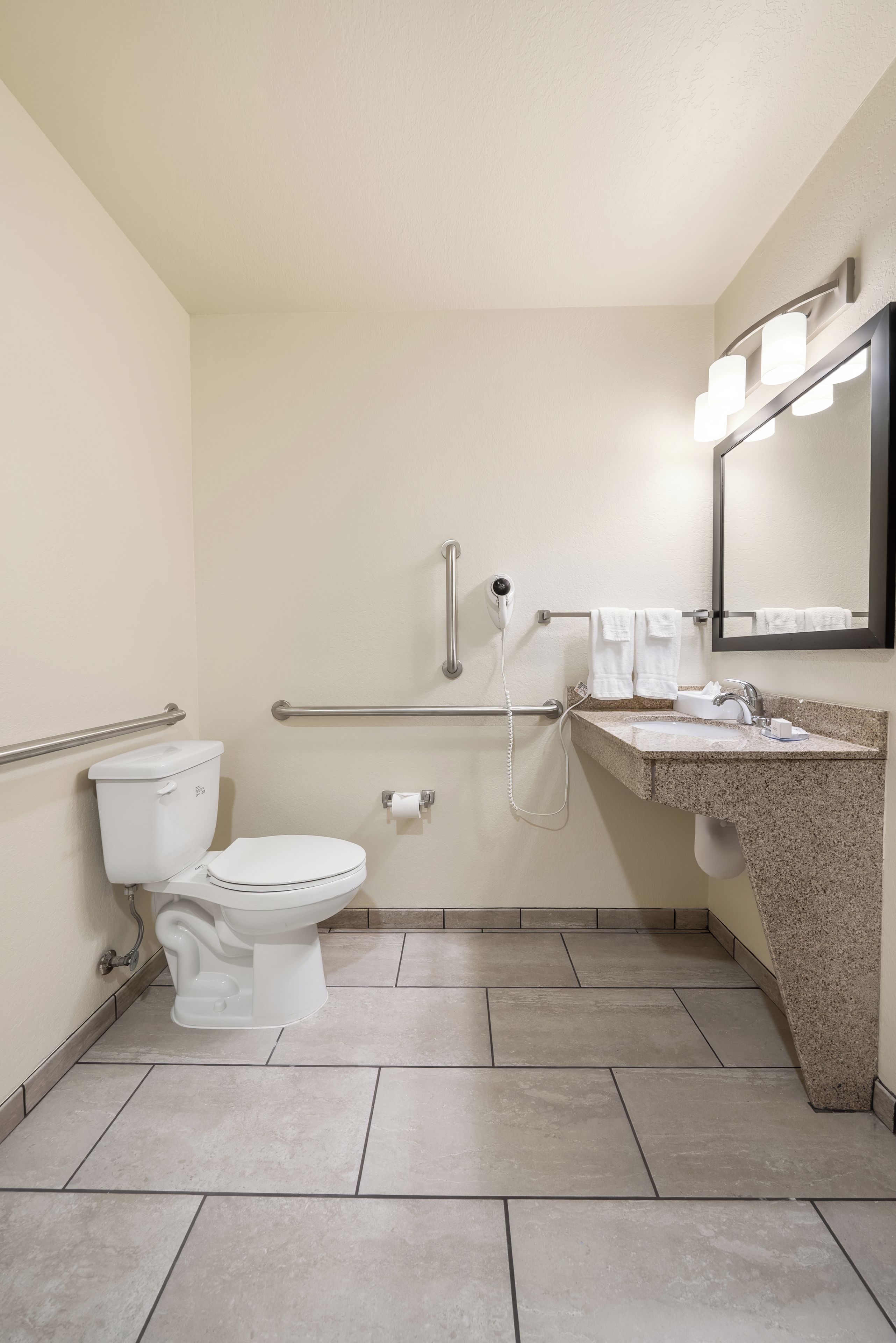 A bathroom with a toilet, grab bars, a hand dryer, a sink with a mirror, and a towel rack.