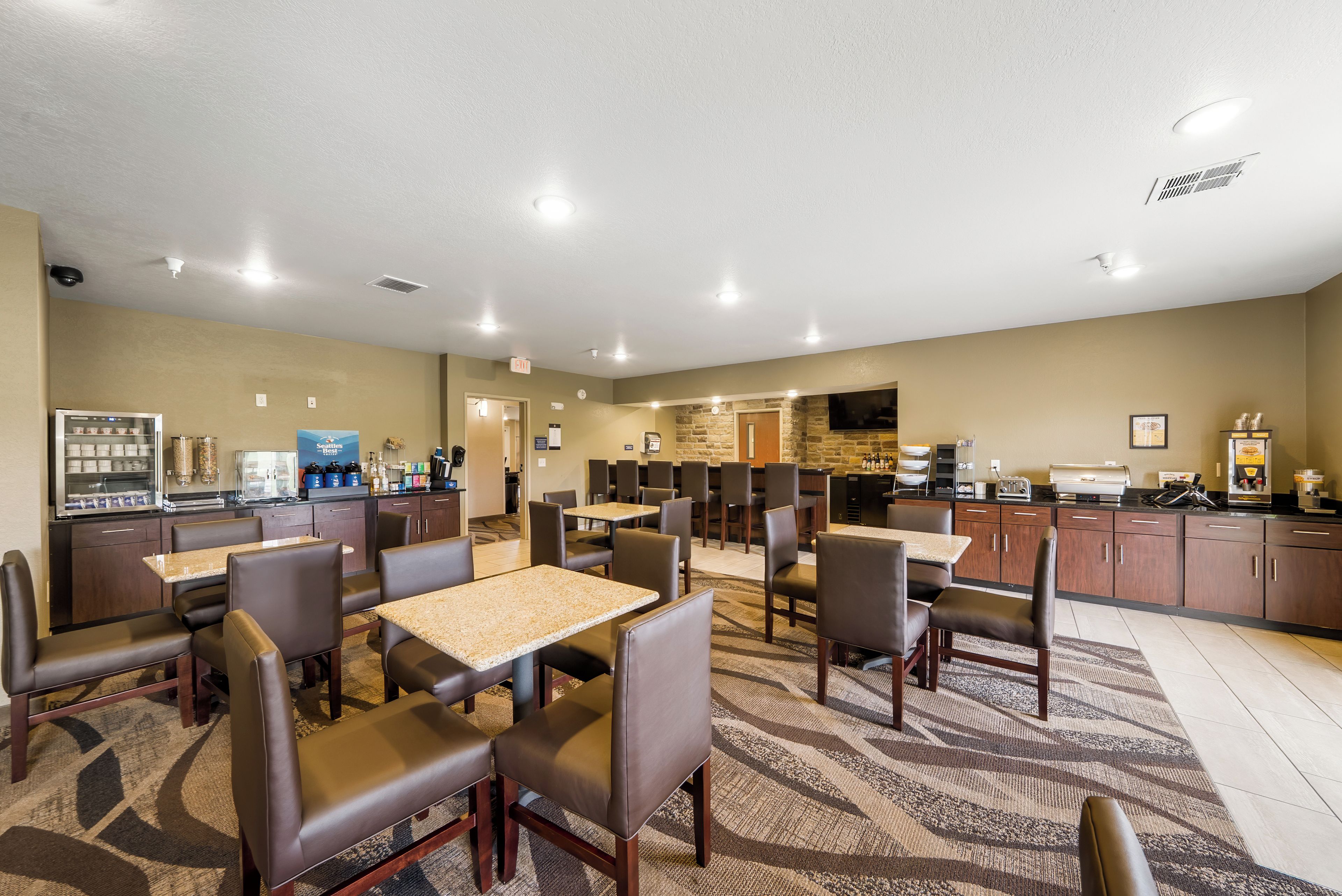 A hotel dining area with multiple tables and chairs, a kitchen area, and a restroom visible in the background.