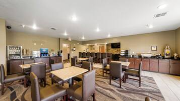 A hotel dining area with multiple tables and chairs, a kitchen area, and a restroom visible in the background.
