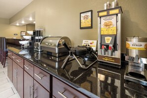 A self-service breakfast area with waffle batter dispensers, syrup dispensers, and a waffle iron.