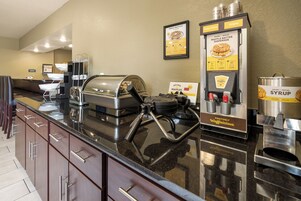 Uma área de café da manhã com serviço por si só, com dosadores de massa para waffles, dosadores de calda e uma máquina de waffles.