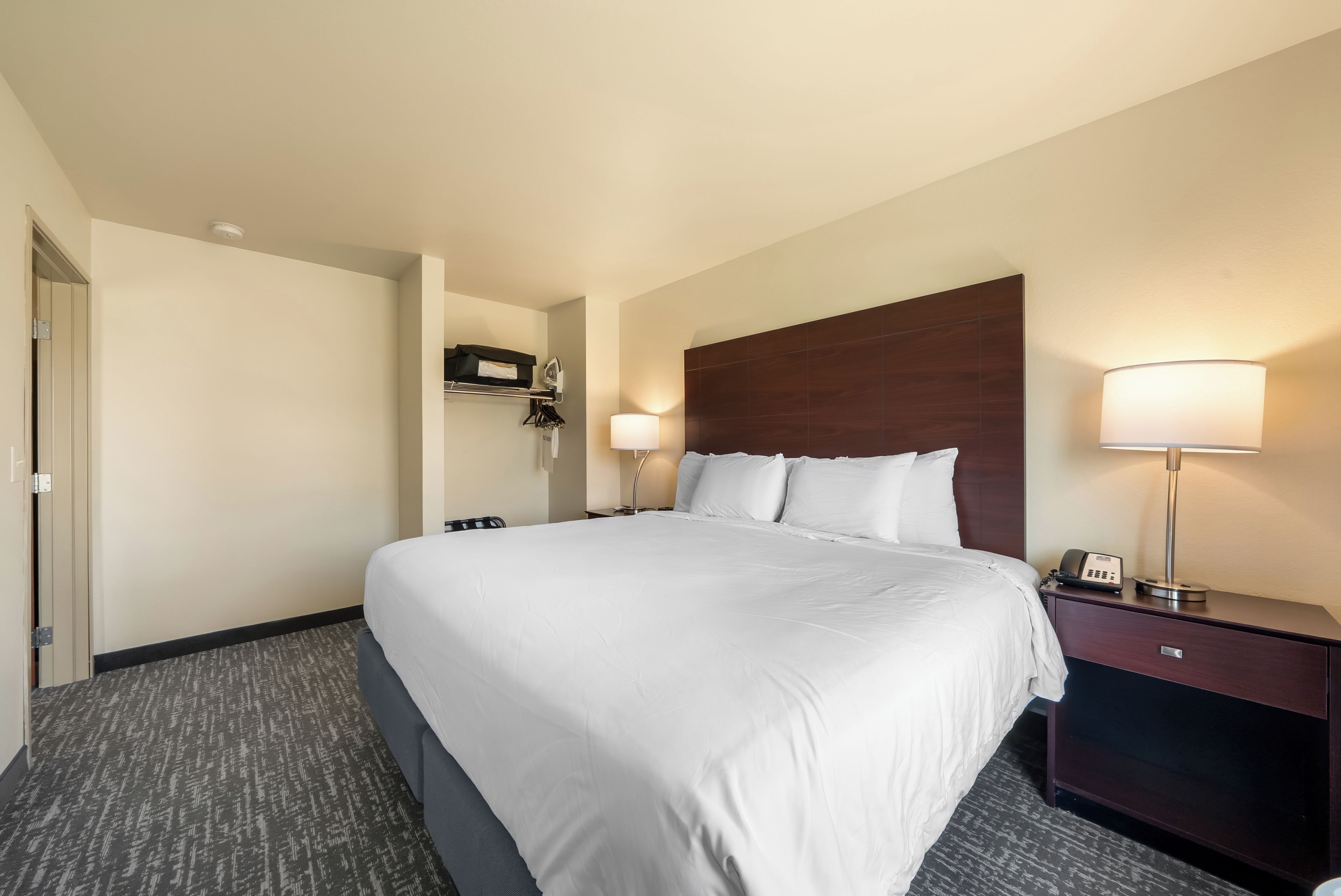 A hotel room with a large bed, two bedside tables, a nightstand, a lamp, and a wall-mounted shelf with a telephone.