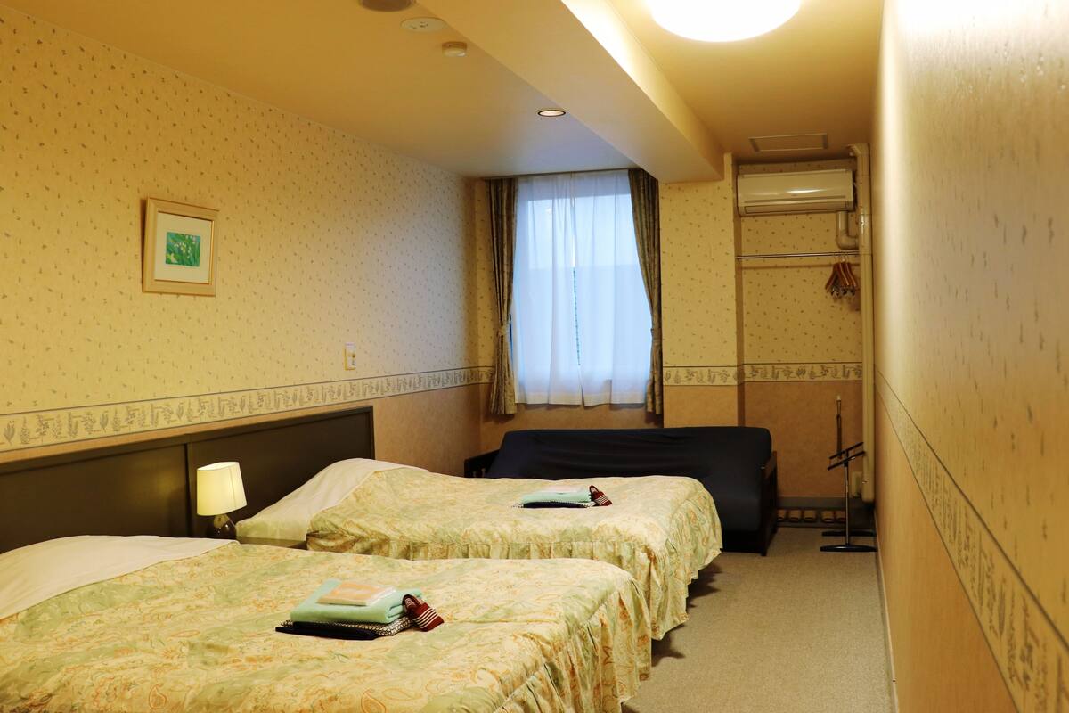 A hotel room with two beds, a window with curtains, a wall-mounted air conditioner, and a small table with a lamp.