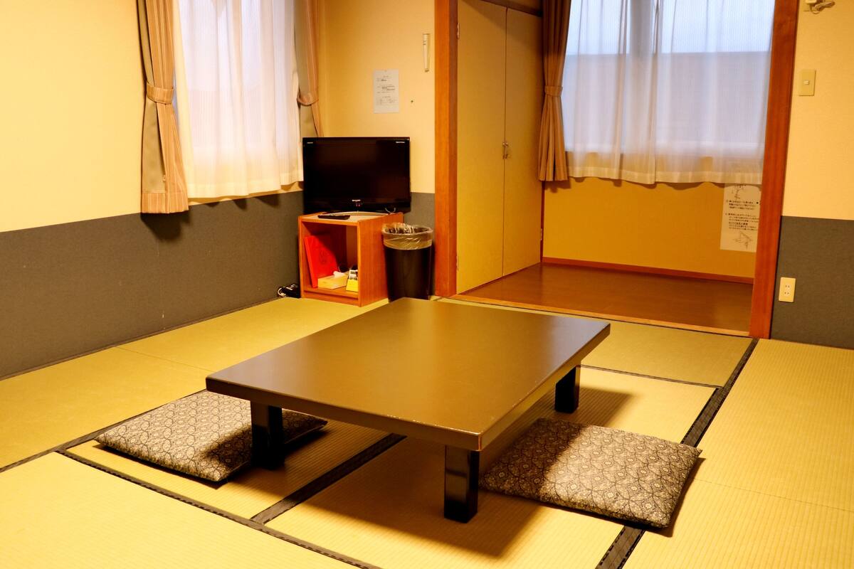 A tatami mat room with a low table, cushions, a TV, and a trash bin.