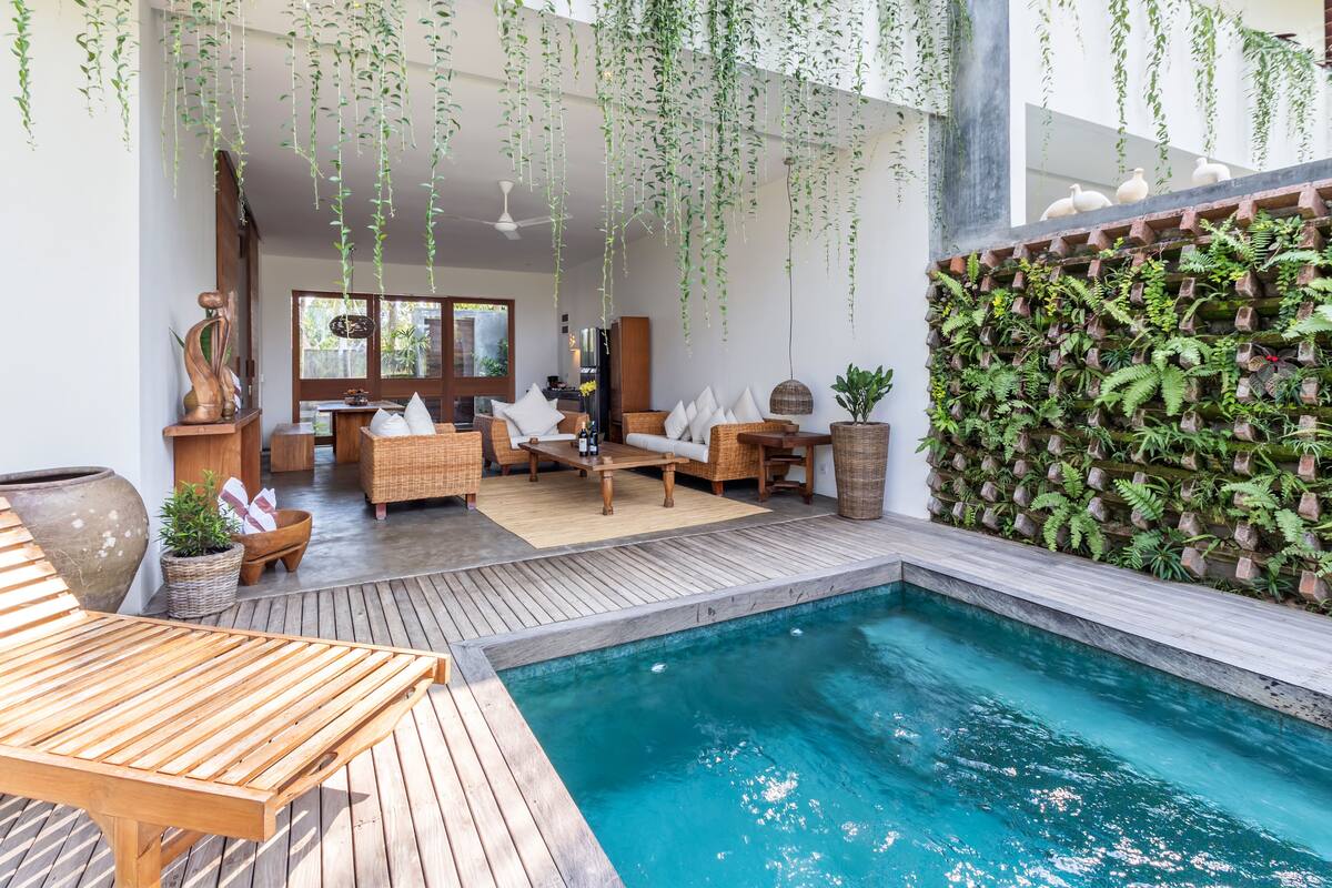A poolside area with a wooden deck, a seating area with wicker furniture, and a wall of hanging plants.