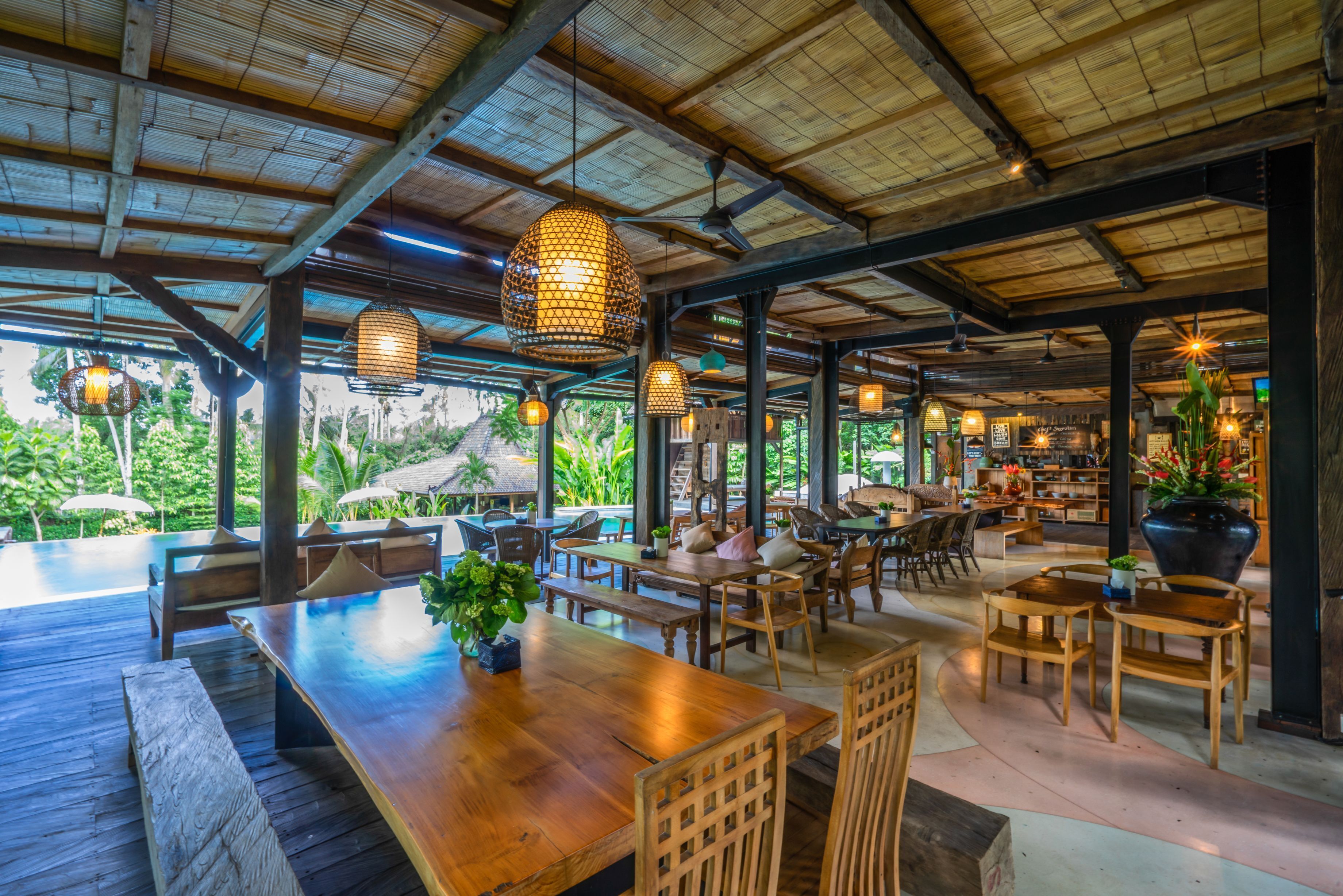 An indoor dining area with wooden tables and chairs, hanging lights, and a view of lush greenery outside.