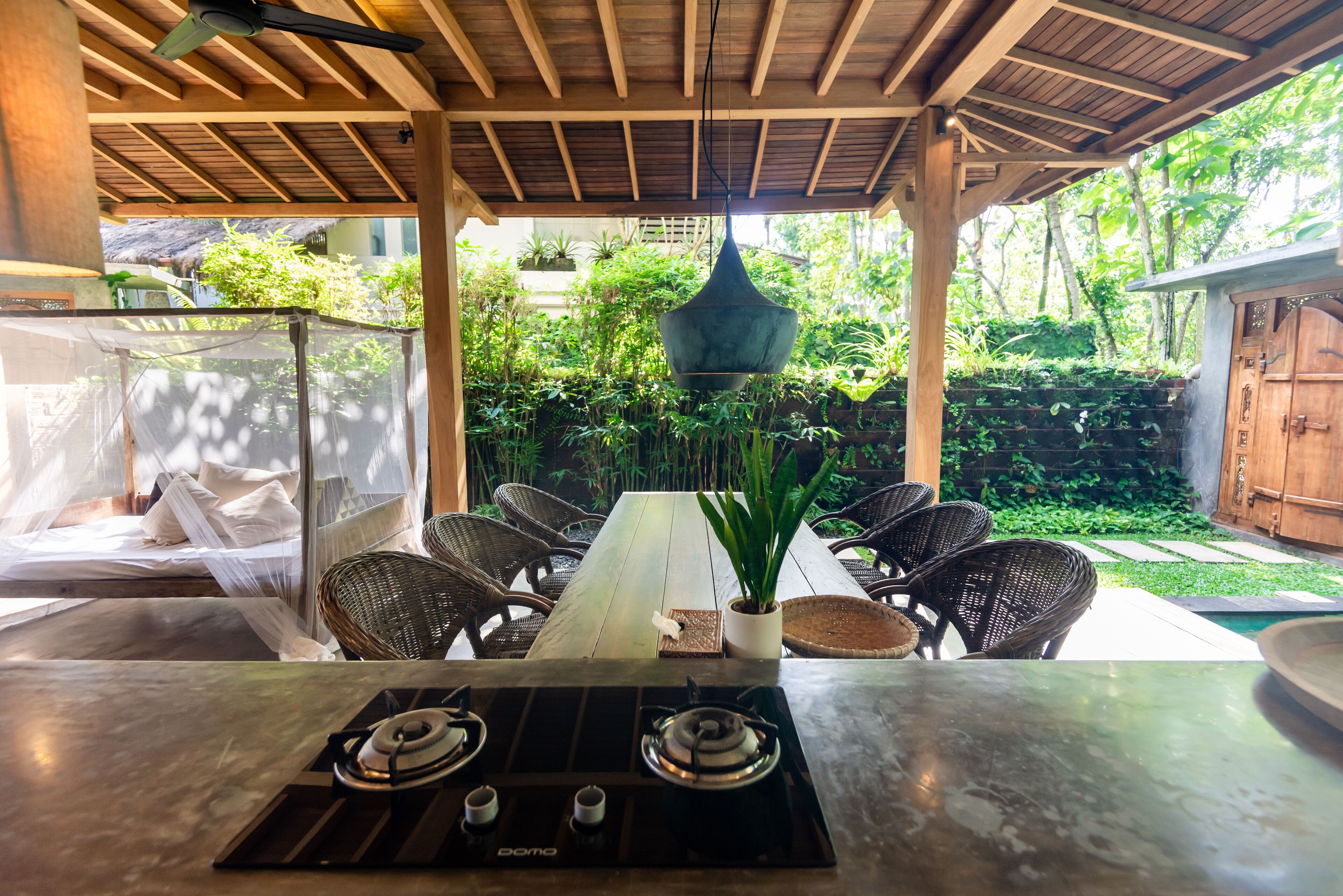 A covered outdoor dining area with a gas stove, wicker chairs, and a potted plant.