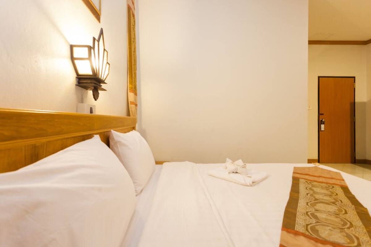 A hotel room with a neatly made bed, a wooden headboard, a wall-mounted light fixture, and a door in the background.