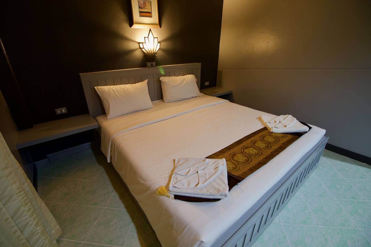 A neatly made bed with white linens and a decorative throw, a bedside table with a lamp, and a wall-mounted light fixture.