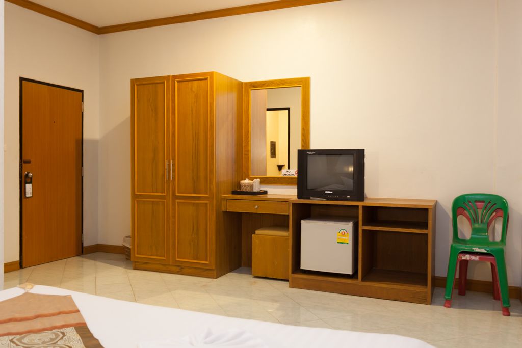 A room with a wooden wardrobe, a small desk with a television, a refrigerator, and a green plastic chair.