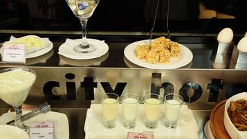 A buffet setup with various food items and beverages, including a martini glass, plates of butter, cream, and yogurt, and shot glasses filled with a creamy beverage.