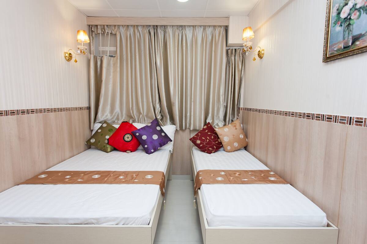 Una habitación de hotel con dos camas individuales, cortinas color beige y lámparas fijadas a la pared.