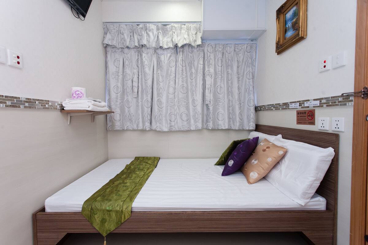 Una cama individual con una manta verde, ropa de cama blanca y cortinas estampadas. Hay un estante con toallas y un cuadro enmarcado en la pared.