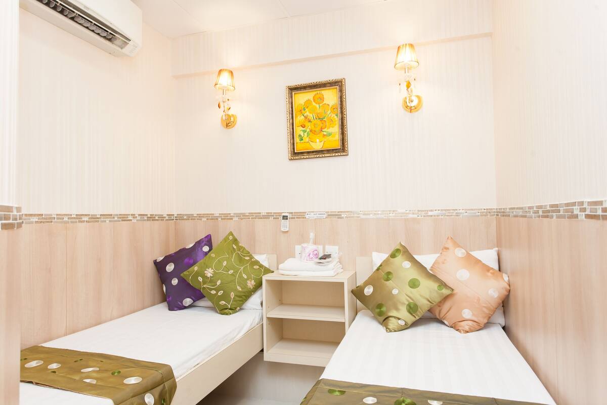 Habitación de hotel con dos camas individuales, una mesita de noche con lámpara, un cuadro en la pared y un equipo de aire acondicionado.