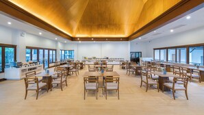 Uma sala de jantar ampla, com várias mesas e cadeiras, um balcão central com vitrine e grandes janelas que proporcionam luz natural.