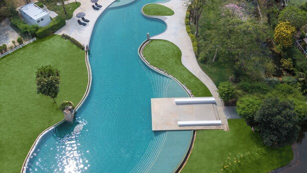 Vista aérea de um resort com uma piscina grande e sinuosa, cercada por gramados bem cuidados, e um hotel com vários andares e telhado de telhas vermelhas.