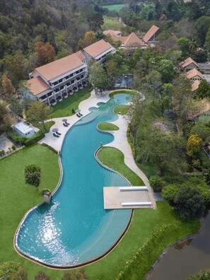 An aerial view of a resort with a large, winding pool surrounded by manicured lawns and a hotel with multiple floors and a red-tiled roof.