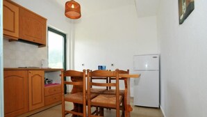 A compact kitchen with wooden cabinets, a dining table with chairs, a hanging light fixture, and a white refrigerator. - Kantaros Apartments (Corfu)