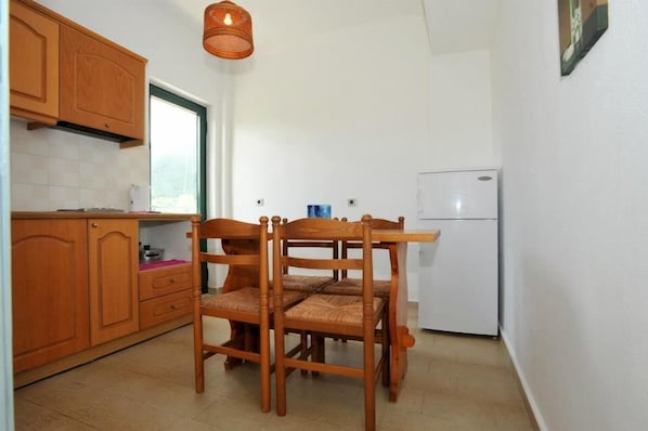 A compact kitchen with wooden cabinets, a dining table with chairs, a hanging light fixture, and a white refrigerator.