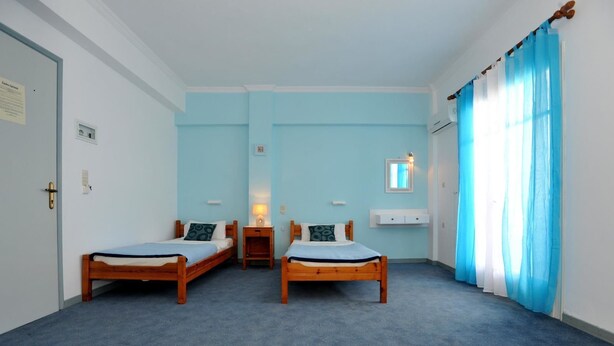 A room with two wooden beds, a small bedside table with a lamp, a white cabinet, and a window with blue curtains. - Kantaros Apartments (Corfu)