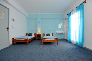 A room with two wooden beds, a small bedside table with a lamp, a white cabinet, and a window with blue curtains. - Kantaros Apartments (Corfu)