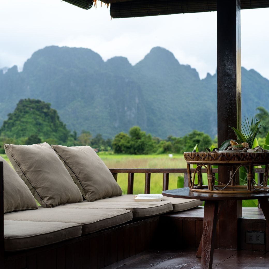 A wooden deck with a cushioned seating area, a woven table, and a view of green mountains.