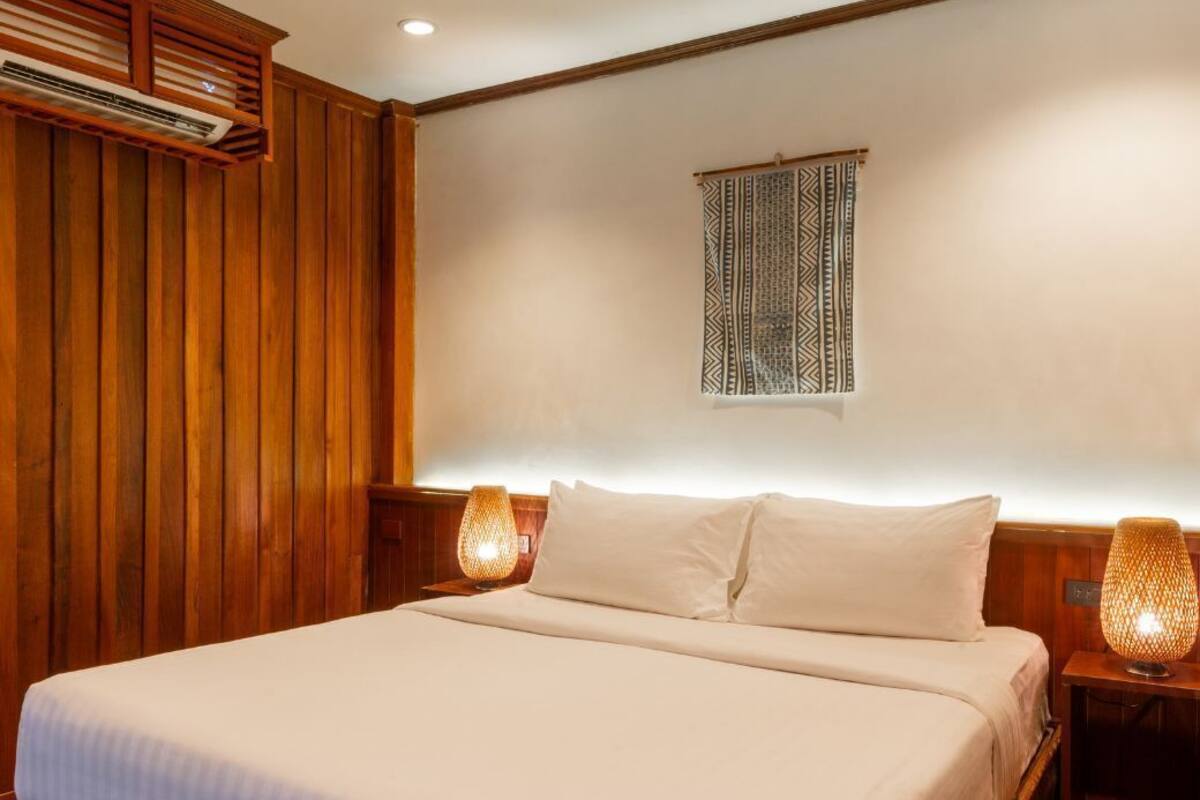 A bedroom with a wooden panel wall, a bed with white bedding, two bedside lamps, and a wall-mounted air conditioning unit.