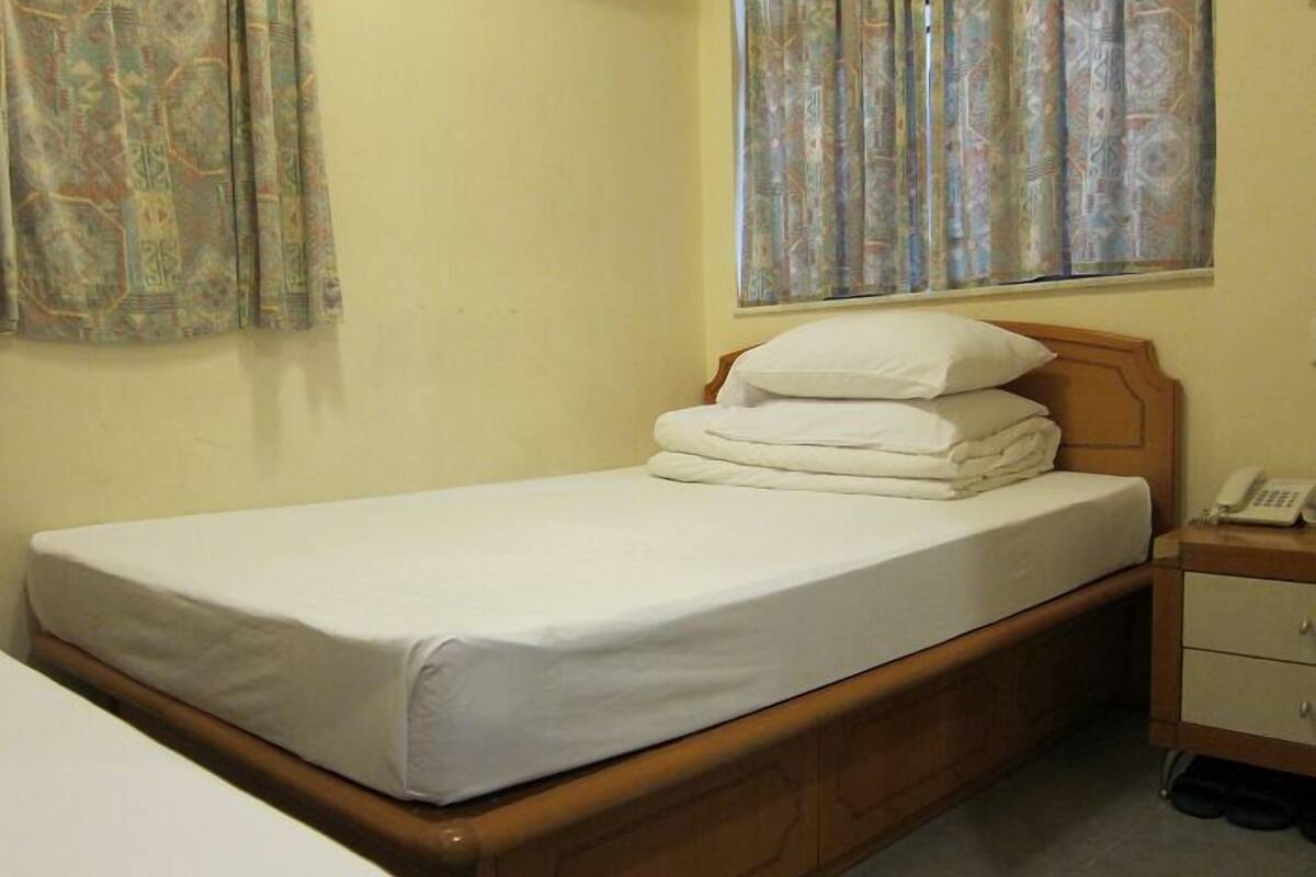 A single bed with a wooden headboard and a white mattress, a bedside table with a phone, and a window with patterned curtains.