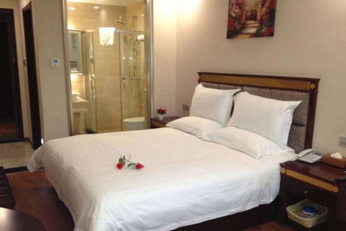 A hotel room with a large bed, a bedside table with a phone and tissues, a small trash bin, and a shower area visible through a glass door.