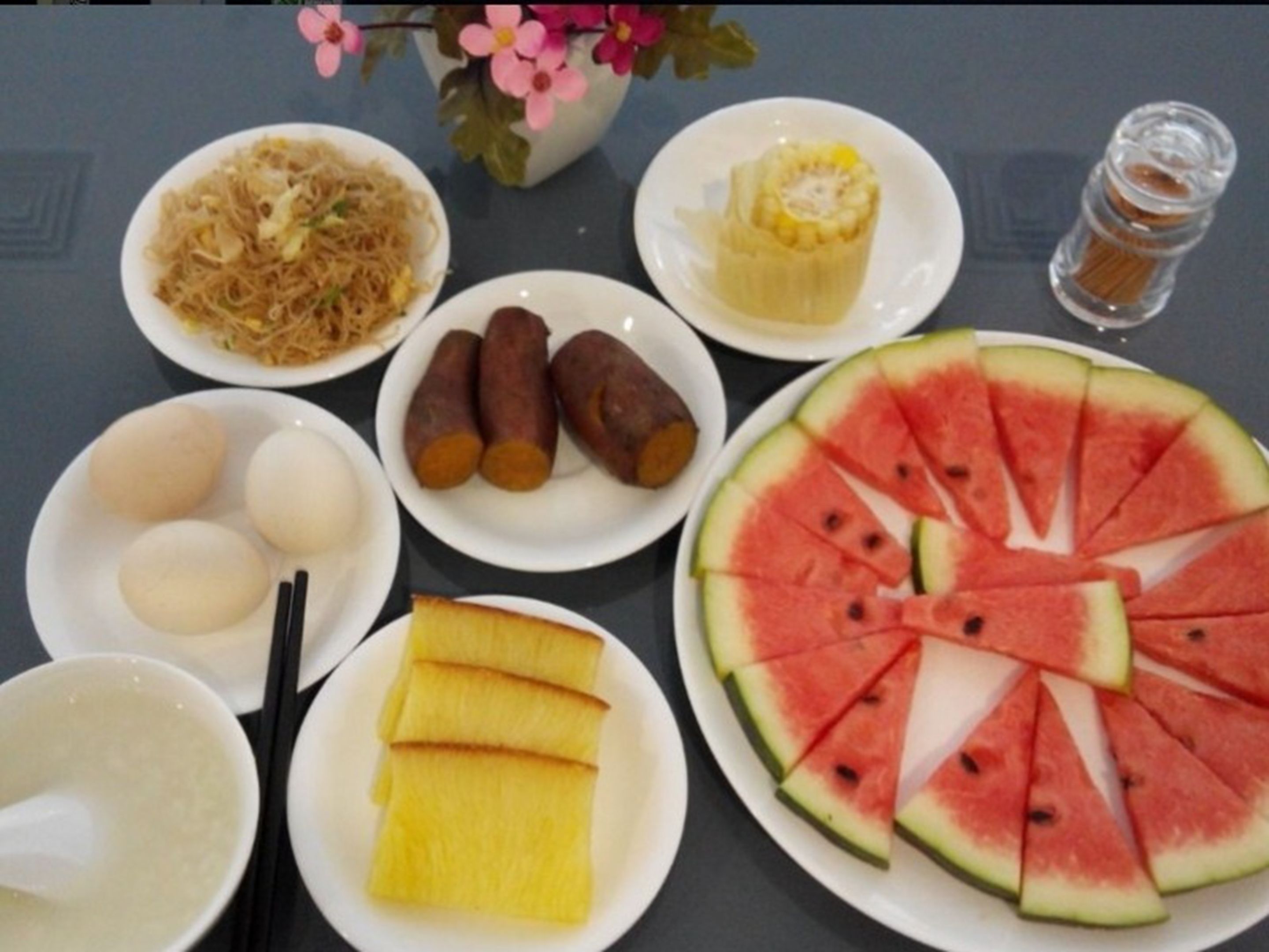 A table with various dishes including noodles, sweet potatoes, eggs, pineapple slices, and watermelon.