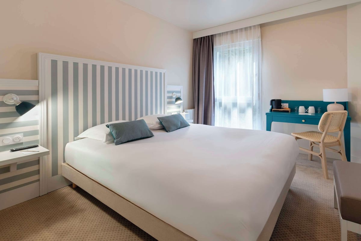 Een hotelkamer met een groot bed, een bureau met lamp, een stoel en uitzicht op het omliggende landschap door het raam.