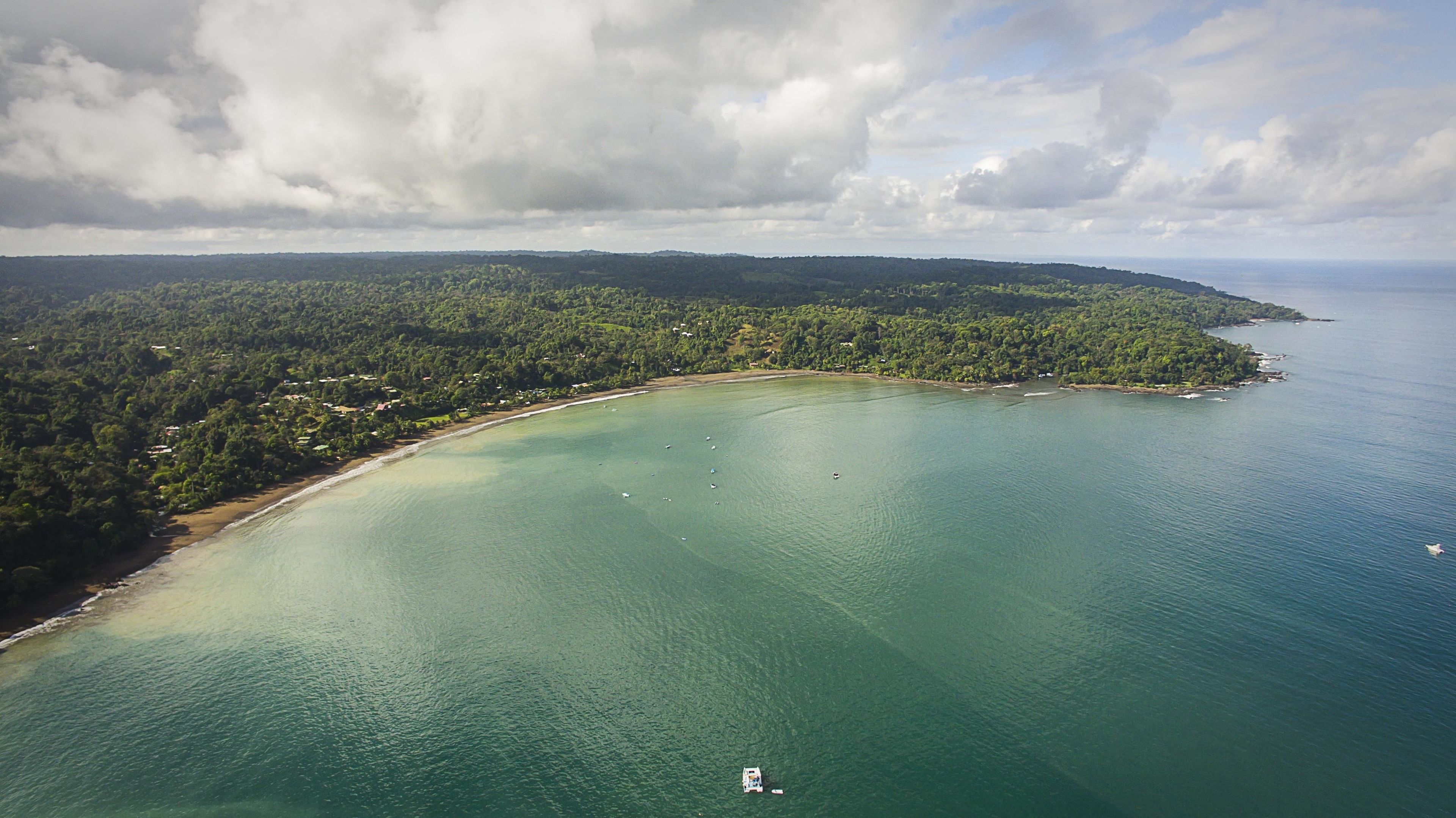 An aerial view of a coastal area with a beach, clear turquoise waters, and a forested peninsula.