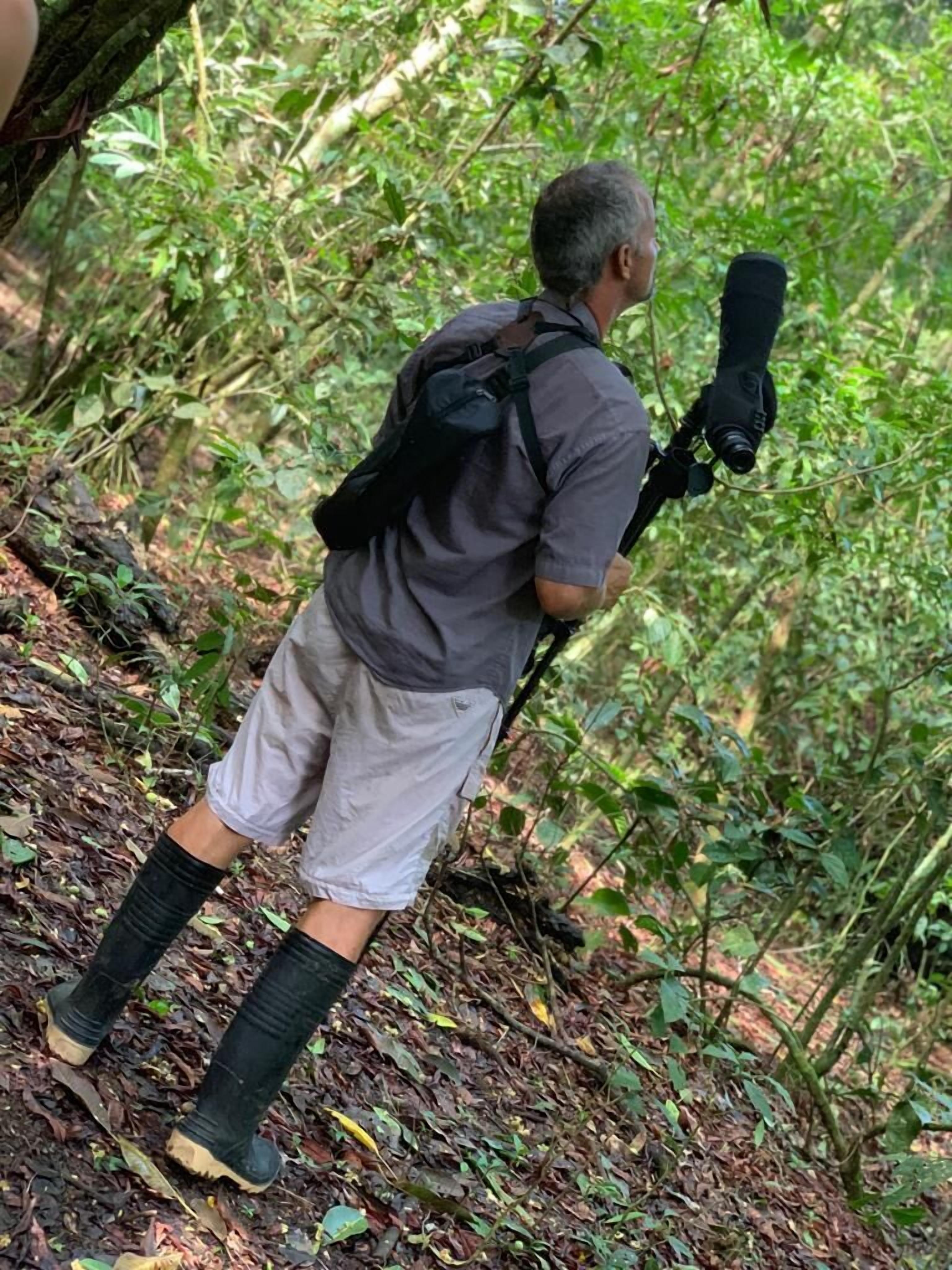 Une personne se trouve dans une zone boisée, porte un sac à dos et tient un appareil photo.