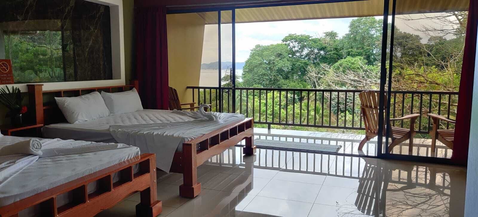 A room with two beds, a balcony with a view of greenery and mountains, and a sign indicating no smoking.