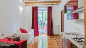 A compact living space with a white sofa, red cushions, a small table with a lamp, and a kitchen area with a stove and sink. - Lh Royal Suites (Rome)