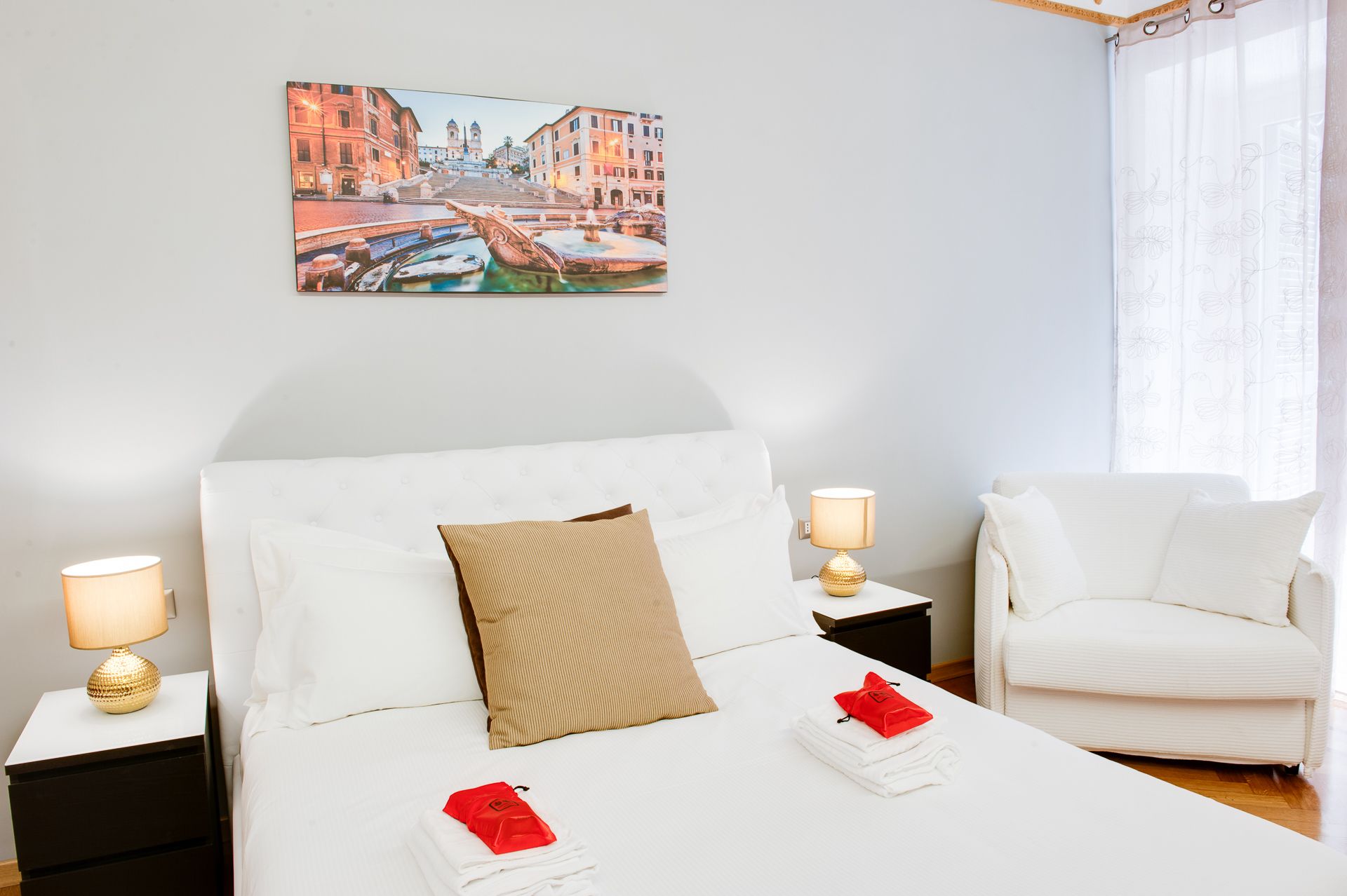 A hotel room with a bed, two bedside tables with lamps, a chair, a window with curtains, and a painting of a cityscape on the wall.