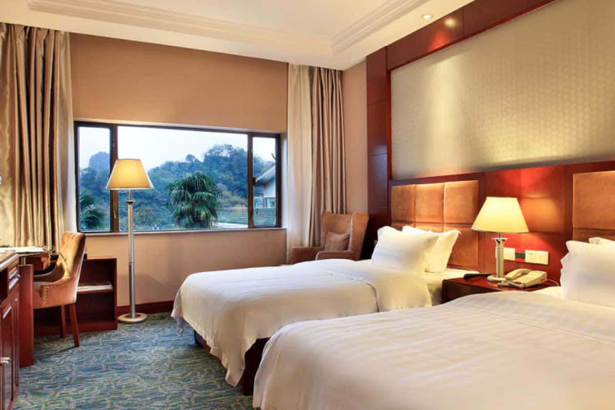 A hotel room with two beds, a desk, a chair, a lamp, and a view of the outdoors.