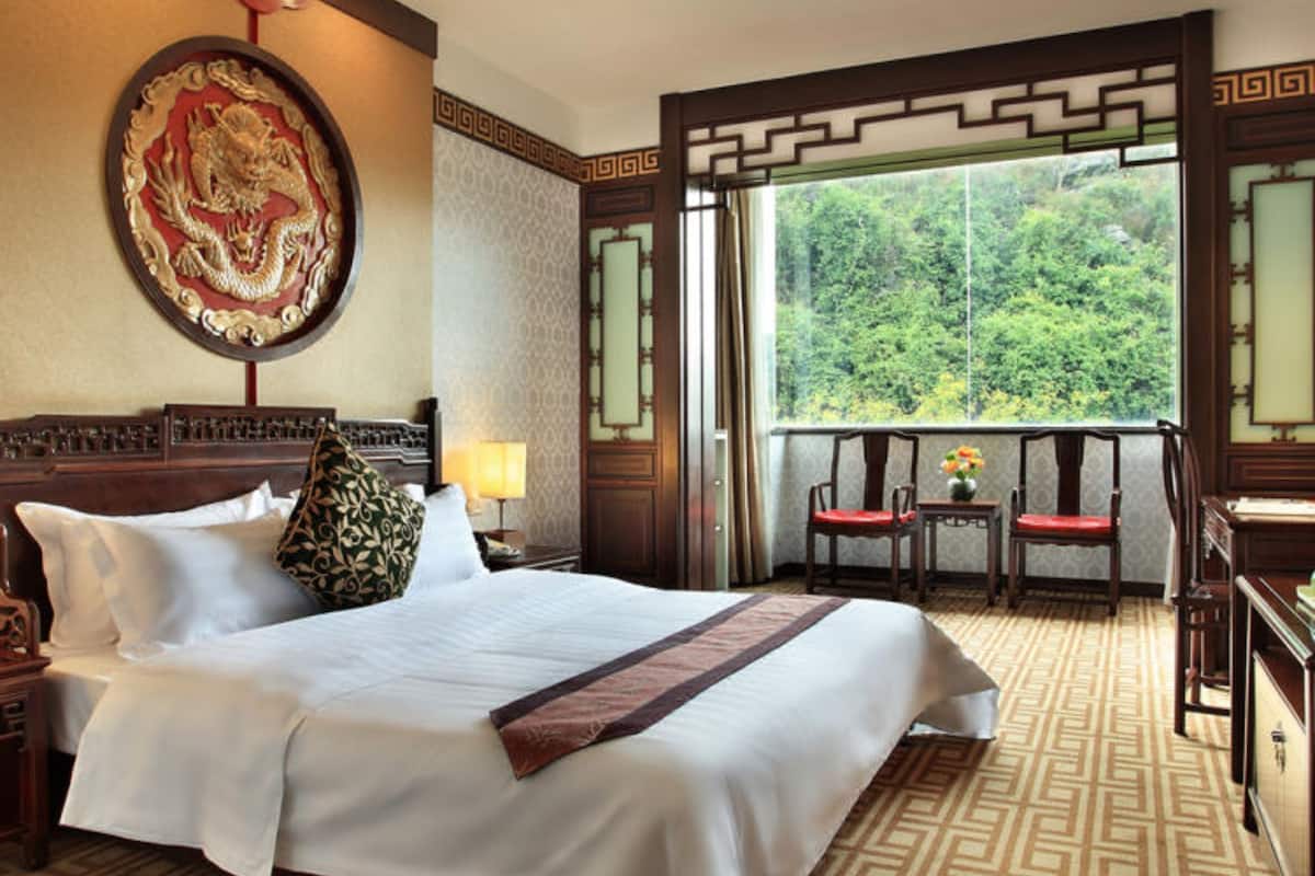 A hotel room with a large bed, a decorative wall hanging, a window with a view of greenery, and traditional wooden furniture.
